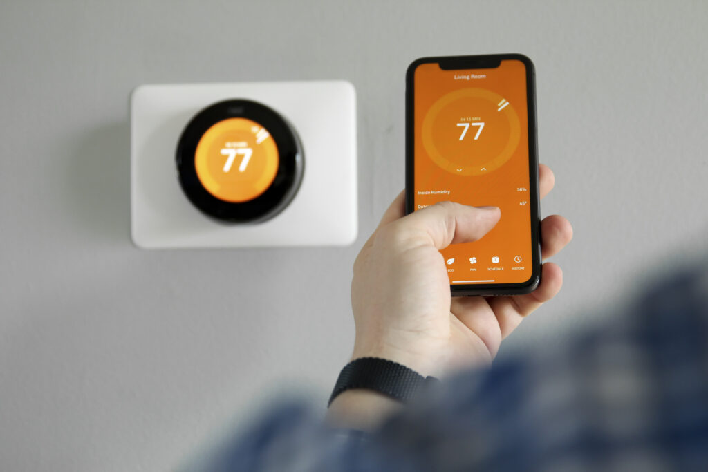 View of person's hand adjusting temperature of thermostat on an app; thermostat on the wall in the background. Display shows 77 degrees.